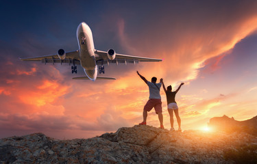 Passenger airplane and happy people on the mountain against colorful sky at sunset. Landscape with commercial airplane and standing man and woman with raised up arms in summer. Couple and aircraft