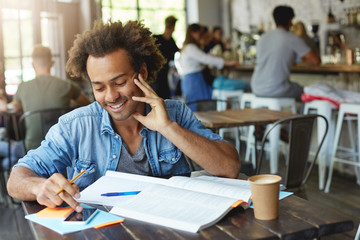 Wall Mural - Waist up portrait of positive smiling Afro American college student typing text message on his smart phone with copy space blank screen while sitting at canteen and working on home assignment