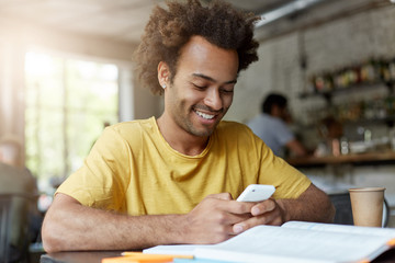 Wall Mural - Cheerful handsome young African American male student in yellow t-shirt browsing internet on smart phone, having rest at coffee shop after classes at university, looking at screen and smiling broadly