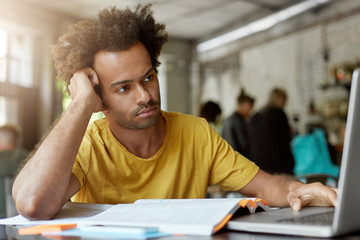 Wall Mural - People, education, modern technologise and youth concept. Dark-skinned mixed race male with curly hair being focused in screen of laptop having thoughtful look leaning at his hand sitting in cafe