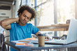 © wayhome.studio  - Serious Afro American male student in blue T-shirt sitting at cafeteria drinking takeaway coffee working at his project using books and laptop touching his neck with hand having pain after work