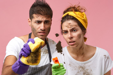 Studio shot of frustrated tired young European wife wearing headband standing close to her bearded husband and staring at dirty spot on window glass. Domestic work and household duties concept