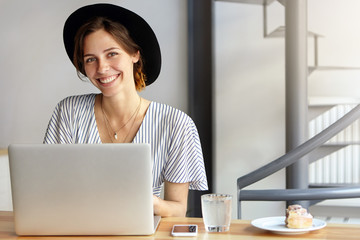 Wall Mural - Businesswoman dressed officially sitting at her working place using laptop computer having pleasant smile rejoicing end of working day. Caucasian female wearing black hat having rest after work