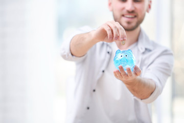 Poster - Young man putting coin into piggy bank on blurred background