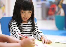 Asian Children Cute Or Kid Girl Smile And Learning For Coloring Or Paint On White Paper With Teacher Or Mother At Nursery Or School On Soft Focus