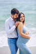 © gorynvd - Courageous and handsome man in a white shirt and blue trousers  hugs her beautiful pregnant wife in a blue dress against the background of the sea.