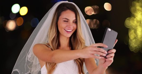 Wall Mural - Close up of cheerful young bride in wedding dress and veil taking a selfie with cell phone outside at night. Happy Caucasian bride making silly faces and photographing herself with smart phone. 4k 
