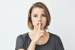 © Cookie Studio - Young beautiful girl in grey t-shirt looking at camera showing keep silence over white background.