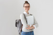 © Damir Khabirov - Indoor photo of college student girl isolated on gray background, smiling at camera, pressing laptop to chest, wearing backpack, ready to go to studies, start new project and suggest new ideas.