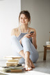 © Cookie Studio - Young beautiful teenage girl surfing internet at phone smiling looking at camera sitting on floor among old books near window over white wall.