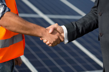 Foreman and businessman shaking hands at solar energy station. Close up view on handshake between worker and man in business suit.