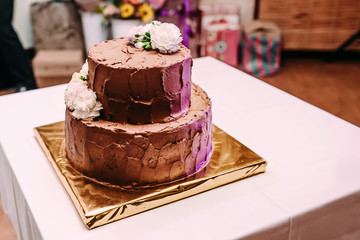 The two-level chocolate wedding cake is decorated with white flowers and stands on the golden stand, on the white table.