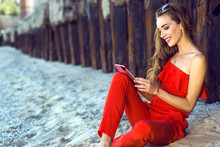 Charming smiling young woman with long hair wearing coral red one shoulder jumpsuit and sunglasses sitting on the beach at the old rusty piles and talking via video connection on her tab. Copy space