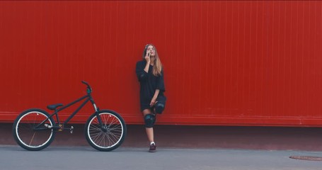 Wall Mural - Outdoor lifestyle portrait of pretty sexy looking young girl in standing near red wall background with street bicycle. Talking on her phone. 4K UHD RAW edited footage