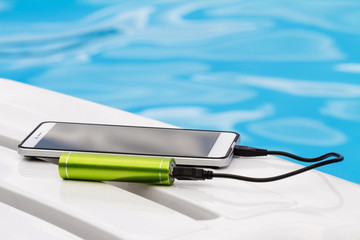 Smartphone connected to the green portable battery charger through USB cable on blue water background. Emergency charging of the phone on a lounger near the swimming pool during the rest.