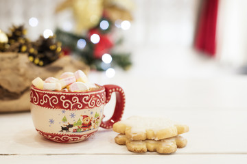 Hot chocolate with marshmallow candies. Christmas cookies shaped in snowflakes,  golden cones and christmass tree lights. White wooden background