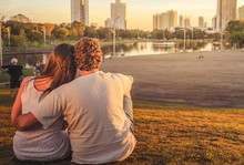 Couple In Love Seated On Grass One Next To Other At Park With A Lake A Nature Around. Sunset With Warm Colors. Couple Aligned To The Left.