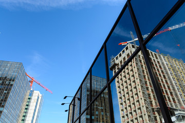 Two concrete buildings under construction with tower cranes at their tops next to a glass building