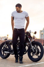 Sporty Biker Handsome Rider Man In White Blank T-shirt Walk Away From Classic Style Cafe Racer Motorbike At Sunset. Vintage Bike Custom Made In Garage. Brutal Urban Lifestyle. Outdoor Portrait.