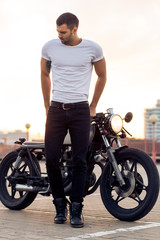 Sporty biker handsome rider man in white blank t-shirt walk away from classic style cafe racer motorbike at sunset. Vintage bike custom made in garage. Brutal urban lifestyle. Outdoor portrait.