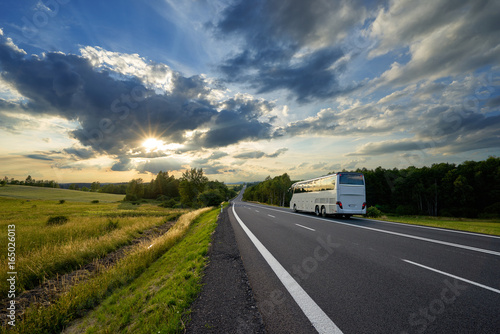 Plakat Autobus podróżuje na asfaltowej drodze w wiejskim krajobrazie przy zmierzchem z dramatycznymi chmurami