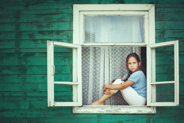 Happy little girl sitting on the windowsill of the old village houses green.