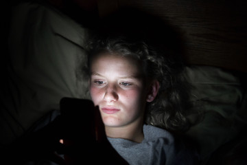 Close up of a teenage girls face lying on a pillow in bed lit up by the light from a cellphone