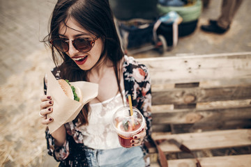 Wall Mural - stylish hipster woman holding juicy burger and lemonade, eating. boho girl biting hamburger, smiling at street food festival. summertime. summer vacation picnic. space for text