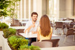© deagreez - Young cheerful guy on a date in cafe outdoors, smiling, talking to his lady and enjoying coffee. Terrace is nice and light, modern, with green plants
