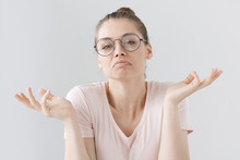 Photo Of European Teenage Girl In Big Round Spectacles Pictured Isolated On Gray Background Pulling Arms Apart In Sign Of Impotence Because She Has No Idea How She Can Cope With Her Problem.
