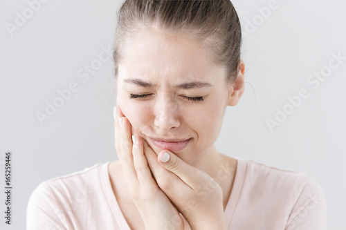 Fotografia Closeup of beautiful teenage girl isolated on gray background touching her face and closing eyes with expression of horrible suffer from health problem and aching tooth, showing dissatisfaction