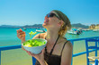 Healthy food. Happy female eating typical Greek salad at outdoor restaurant. Healthy Mediterranean diet concept. Methoni Beach on background in Greece. Woman enjoying healthy food in summer vacation
