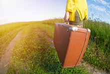 Young Woman Carries Brown Vintage Suitcase In The Field Road During Summer Sunset. Travel Concept.