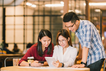 Young Asian college students or coworkers using digital tablet and smartphone together at coffee shop, diverse group. Casual business, freelance work at cafe, social meeting, or education concept