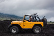 A curly-haired man drinking water is sitting in the offroad yelow vehicle parked at the top of a valley with volcanic rock and mountains in Bali, Indonesia