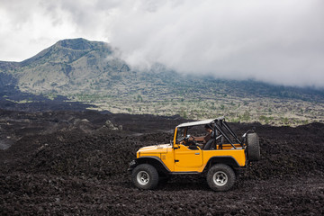 A curly-haired man is sitting in the offroad yelow vehicle parked at the top of a valley with volcanic rock and mountains in Bali, Indonesia