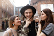 © shurkin_son - Two beautiful stylish young women standing in urban surroundings, hugging handsome bearded hipster in hat, flirting with him as he looking at camera with cheerful smile, feeling pleased and happy