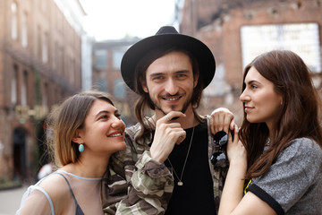 Two beautiful stylish young women standing in urban surroundings, hugging handsome bearded hipster in hat, flirting with him as he looking at camera with cheerful smile, feeling pleased and happy