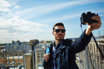 Wall Mural - Portrait of handsome young man taking selfie photo while standing on rooftop viewing platform against panoramic city view and coin-operated binoculars in background