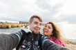 © satura_ - Young laughing couple hiking taking selfie with smart phone. Happy young man and woman taking self portrait with sea or ocean scenery on background. Winter time