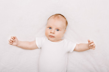 Closeup portrait of adorable funny white Caucasian baby with blue grey eyes lying on bed. Aware cute newborn on white background in studio. Little surprised child looking in camera making faces.