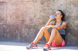 Young smiling female resting after an active training with water bottle while siting on sidewalk, satisfied fit woman resting after an active training, drinking cold water.