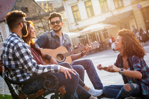 Plakat Grupa ludzi spotyka się na ulicy miasta. Siedzą na ławce, śpiewają i grają na gitarze.