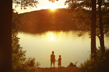 Children At The Water Watching The Setting Sun. Two Boys Holding Hands Standing On The Shore. Back View. Copy Space For Your Text 