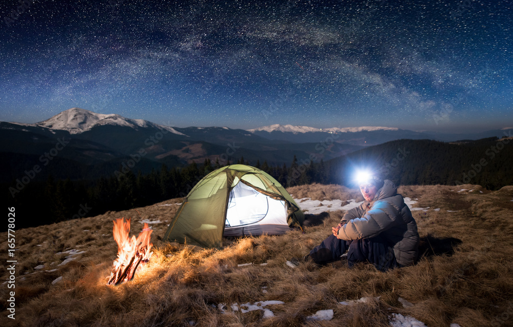 Male tourist have a rest in his camp at night. Man with lighting headlamp sitting near campfire and tent, looking to the camera under beautiful sky full of stars and milky way - obrazy, fototapety, plakaty 