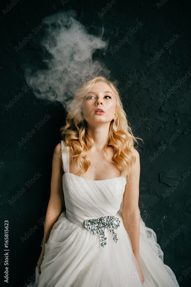 Photo Stock Beuatiful odd blonde bride with curly hairstyle in white  expensive wedding dress smoking electronic cigarette with thin smoke cluds.  Young girl relaxing before marriage. Female on black wall