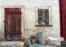 Old House Facade With Rustic Wooden Red Door And Window With Closed Green Shutters And Wooden Chopping Blocks And A White Chair In The Foreground