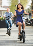 Fototapeta Miasto - Happy young man and woman with electrkc bikes