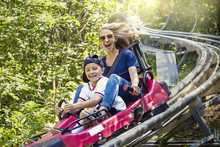 Smiling Women And Her Boy Riding Downhill Together On An Outdoor Roller Coaster On A Warm Summer Day. She Has A Fun Expression As They Enjoy A Thrilling Ride On A Red Amusement Park Ride