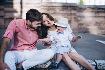 Portrait of young stylish couple sitting on the pavement in old town. Handsome boy happy with his parents. They are smiling together.